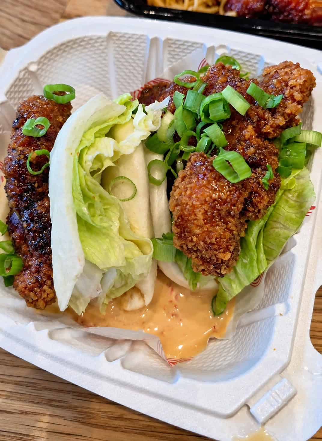Two Crispy Chicken Buns in a white takeout container with soft steamed bao bread, crispy fried chicken, lettuce, and orange sauce