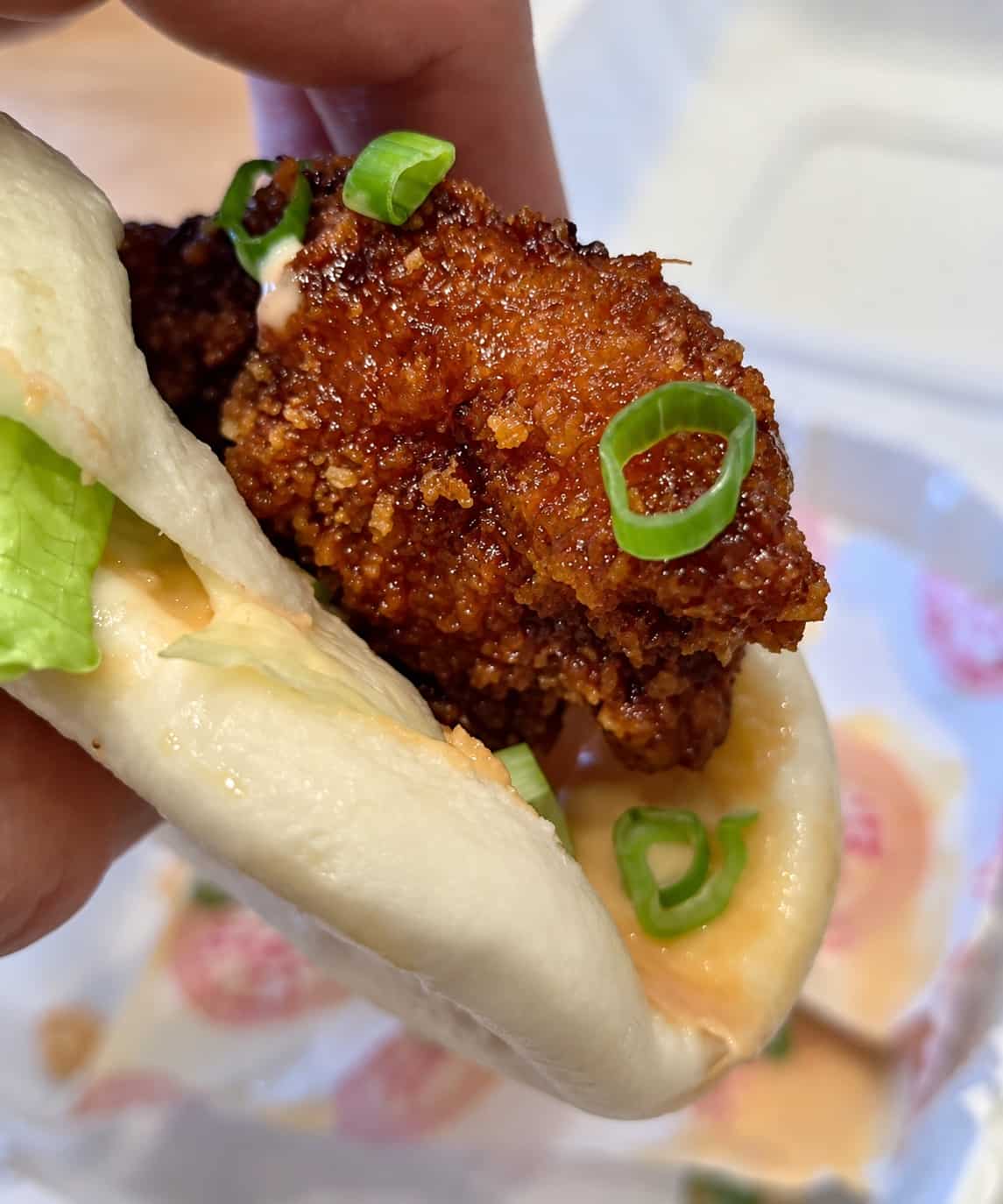 Hand holding a Crispy Chicken Bun showing layers of soft white bao bread, crispy glazed chicken, lettuce, and sauce