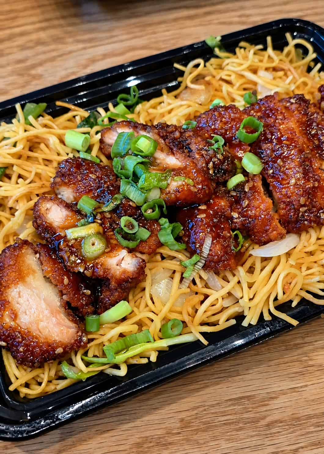 Black tray of pan-fried noodles topped with sliced crispy chicken coated in dark sauce and garnished with green scallions