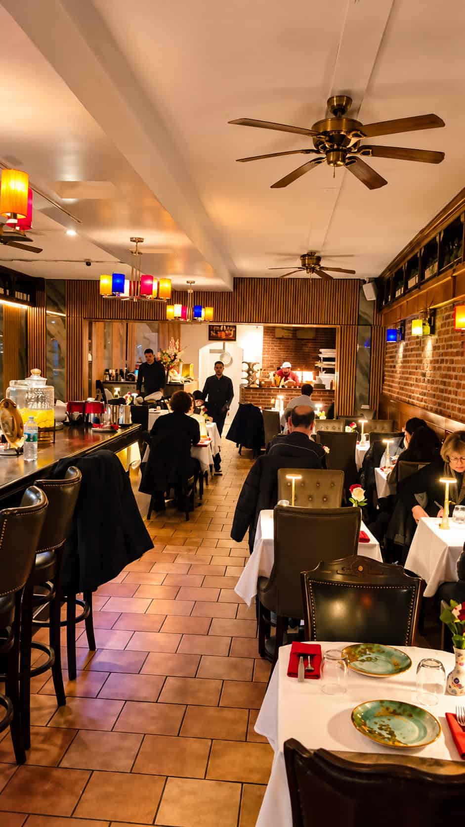 Upscale dining room interior at Ali Baba featuring white tablecloths, exposed brick, dark leather chairs, and colorful ceiling lanterns