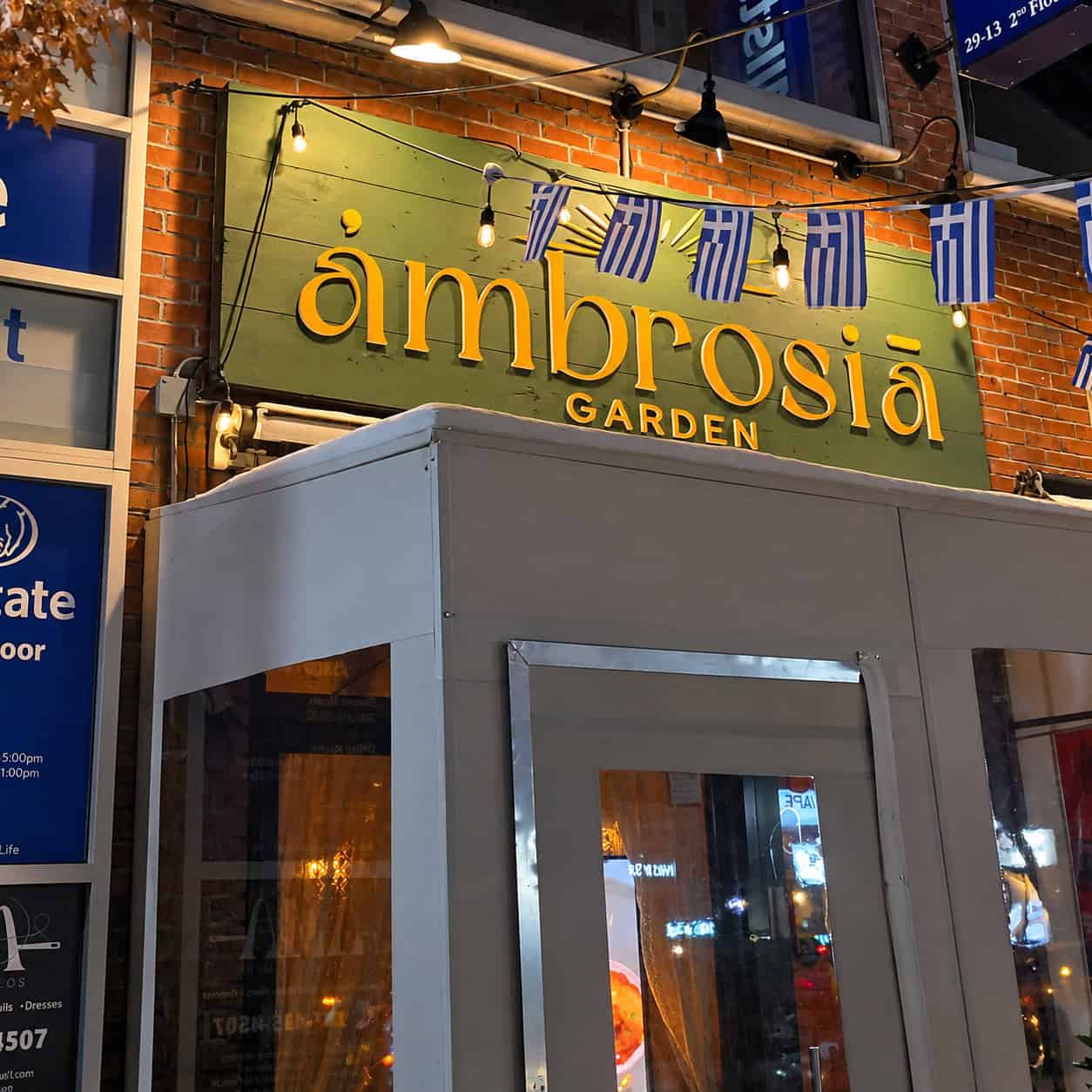 Illuminated green wooden sign and Greek flags at the entrance of Ambrosia Garden restaurant on 23rd Avenue in Astoria