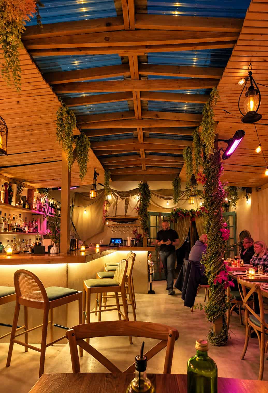 Warm indoor garden dining room and bar at Ambrosia Garden with hanging plants, wooden beams, and cozy lighting