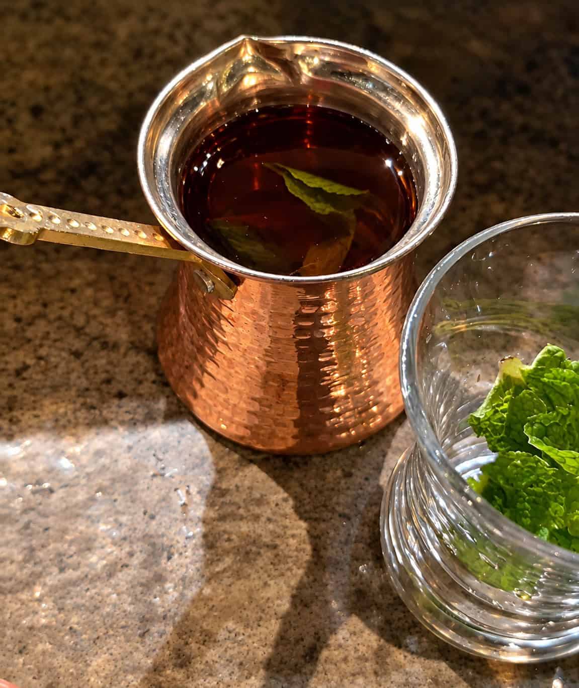Traditional hammered copper pot filled with hot mint tea next to a glass filled with fresh mint leaves