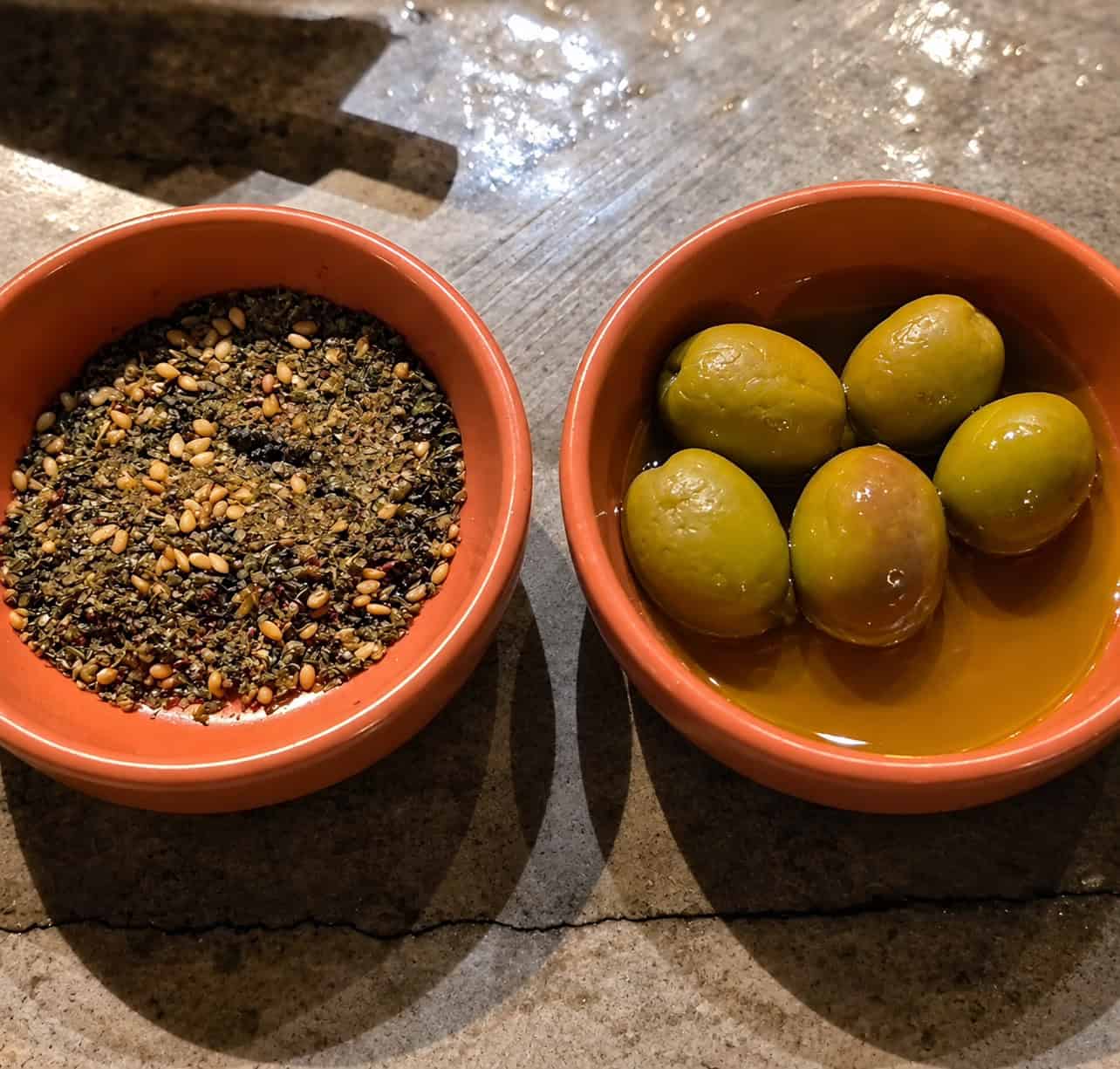 Bowls of green olives bathed in olive oil next to zaatar spice