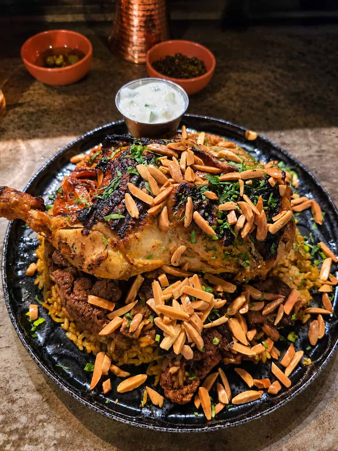 Large platter of Palestinian Maklouba topped with grilled chicken, toasted slivered almonds, and side of yogurt