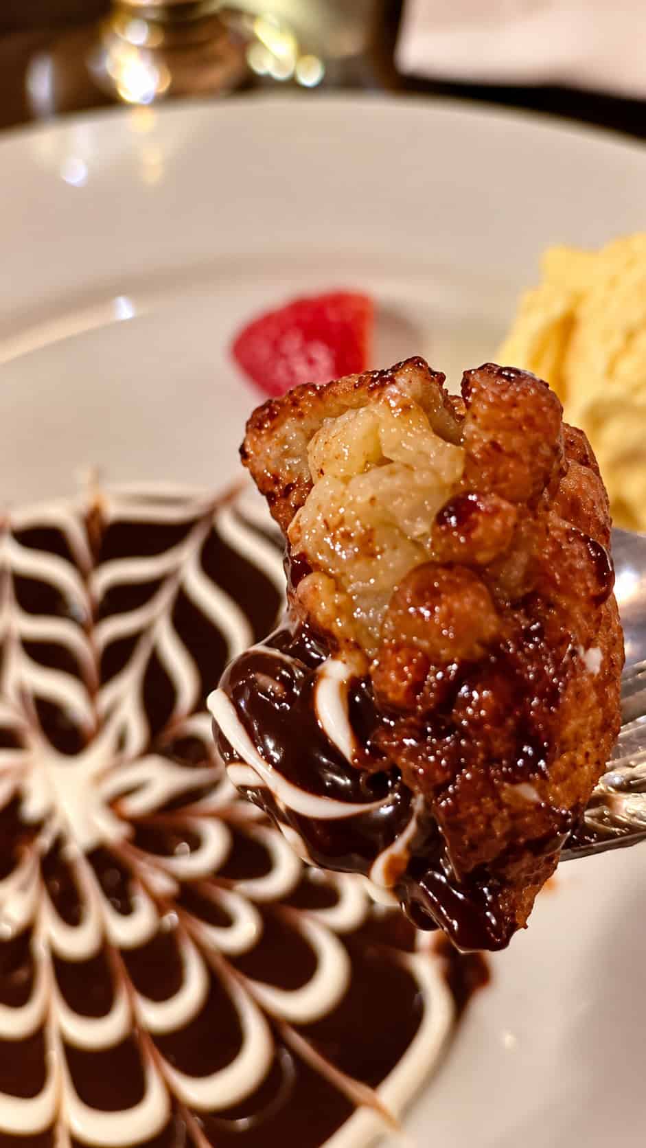 Fork holding a warm fried banana fritter dipped in chocolate sauce over the dessert plate