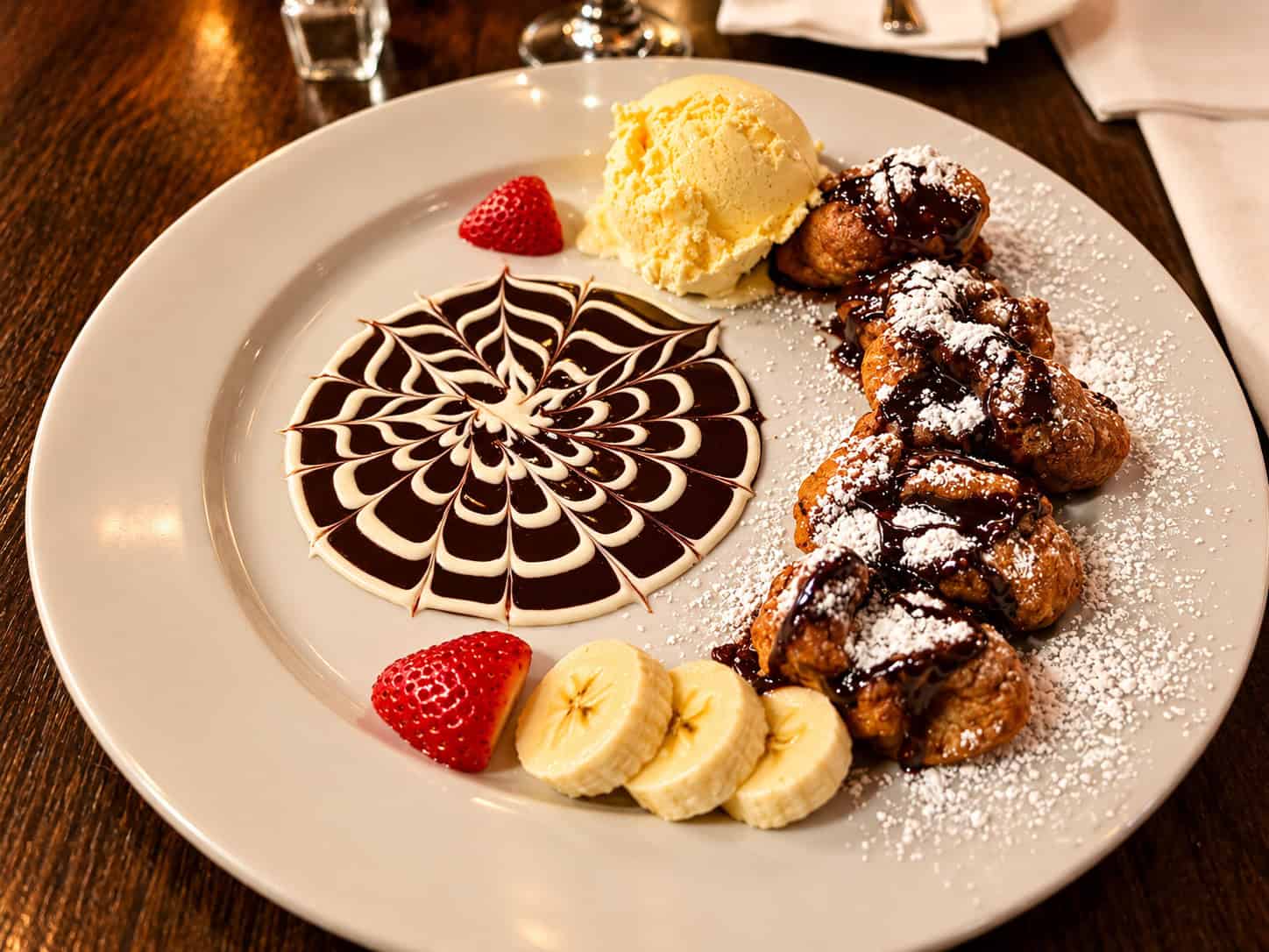 Plated Banana Fritters dessert with chocolate drizzled fritters, banana ice cream, sliced fresh bananas, strawberries, and a spiderweb chocolate sauce design