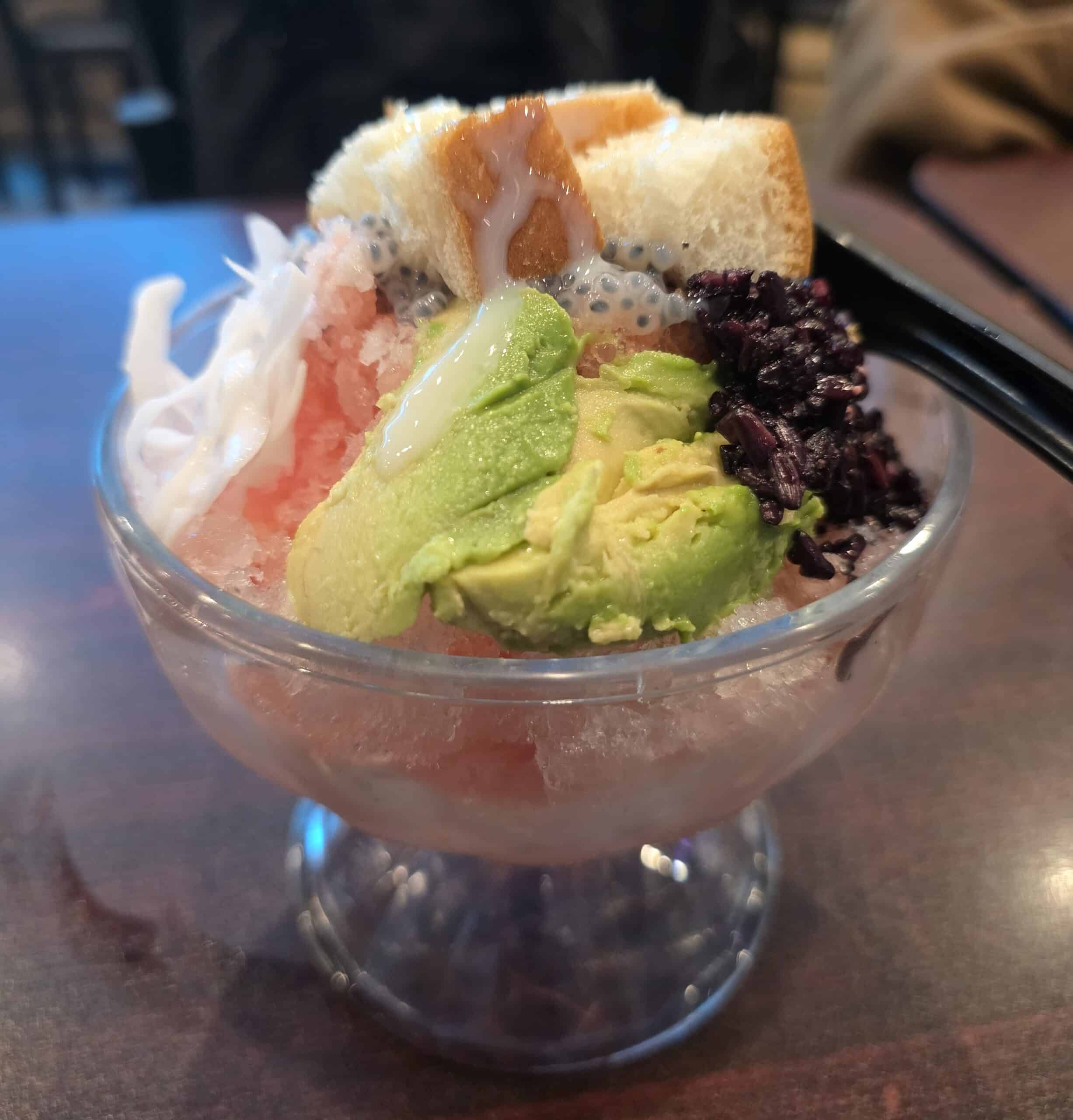 Glass goblet filled with Es Doger Indonesian shaved-ice dessert topped with fresh sliced avocado, young coconut strips, sweet bread cubes, basil seeds, black sticky rice, and condensed milk