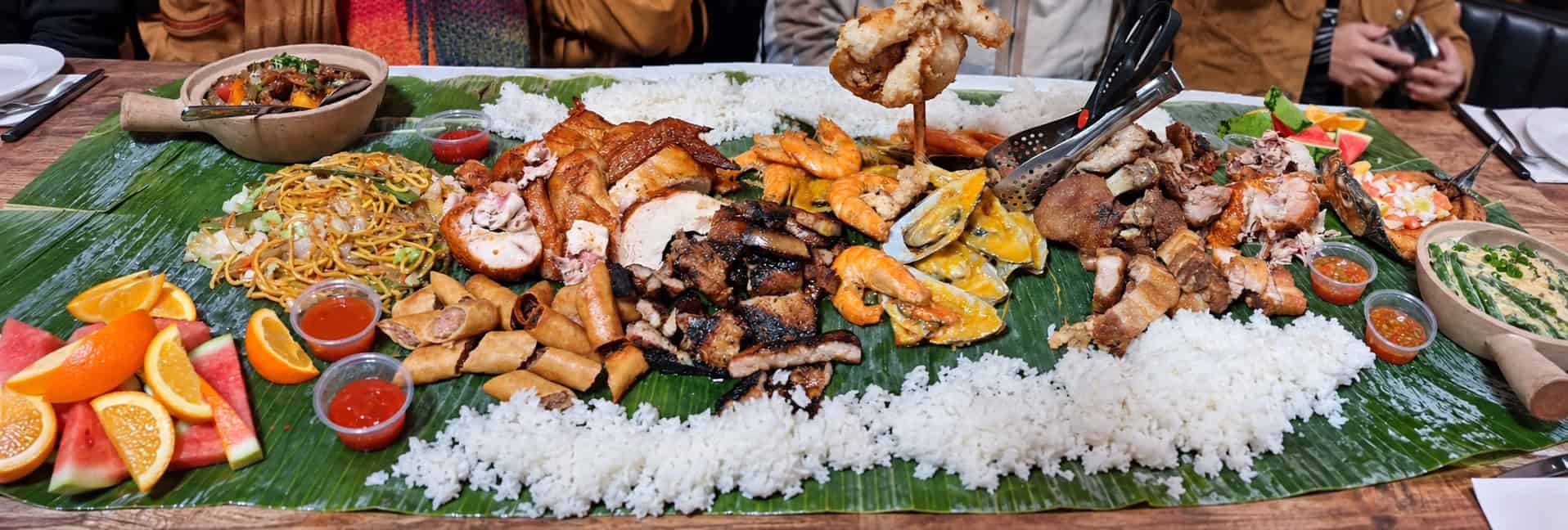 Full overhead view of a massive Filipino kamayan feast spread over banana leaves with rice, grilled meats, lumpia, noodles, and fresh fruit