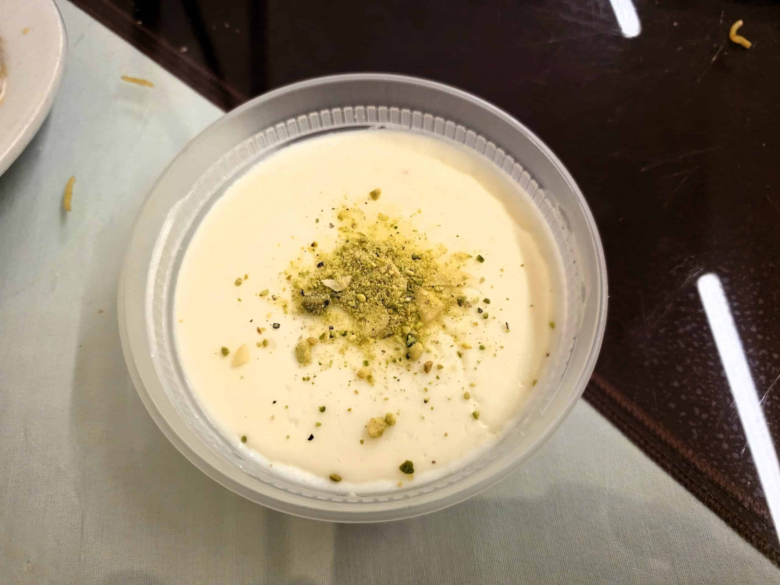 Small plastic container of white Afghan Firnee milk pudding topped with crushed green pistachios