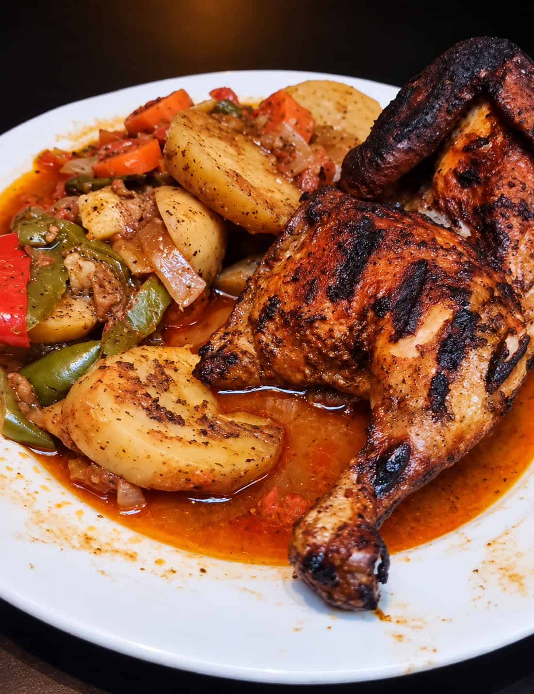 Char-grilled half chicken served on a plate next to stewed potatoes, carrots, and peppers in savory sauce