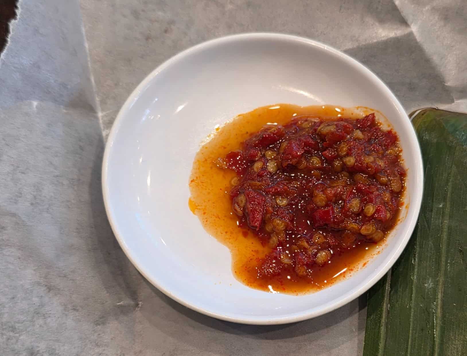 Small white dish of fiery red Indonesian sambal chili sauce alongside banana leaf wrapping