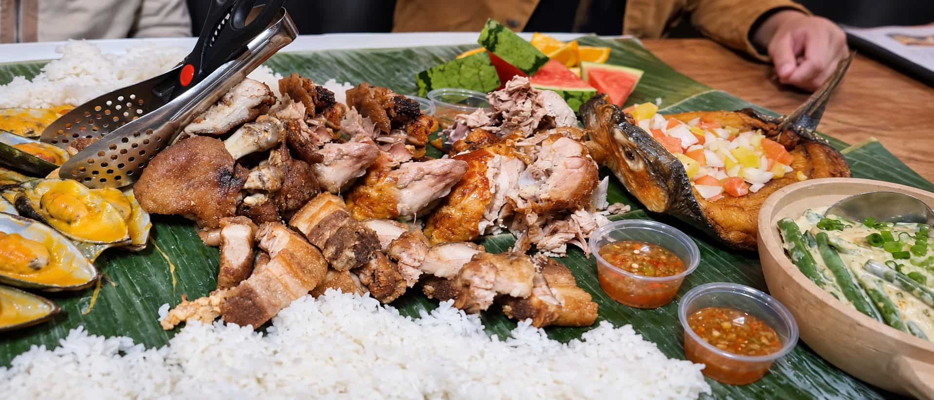 Section of banana-leaf kamayan spread featuring Crispy Pata pork knuckle, grilled chicken, fried milkfish, mussels, and steamed rice