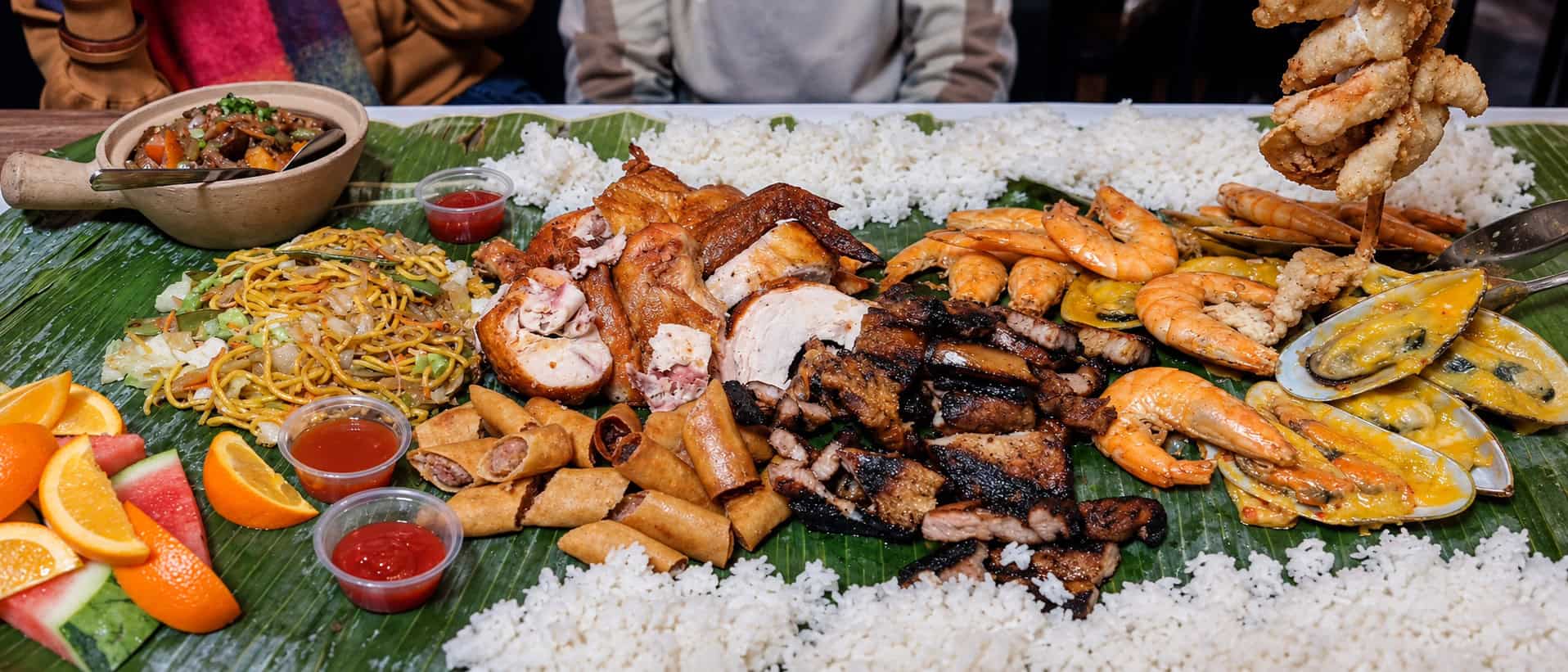 Section of kamayan platter showing Lumpia Shanghai spring rolls, Pancit Bihon noodles, grilled shrimp, and fresh fruit