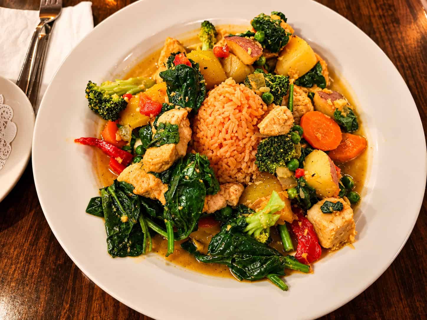 Large plate of Coco Cabana Pollo featuring coconut curry chicken, broccoli, spinach, carrots, and potatoes arranged around a mound of rice
