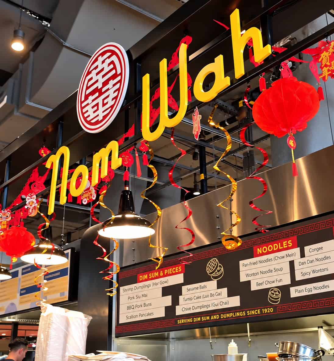 Nom Wah food stall counter inside Market 57 with yellow illuminated sign, red Chinese lanterns, and menu boards