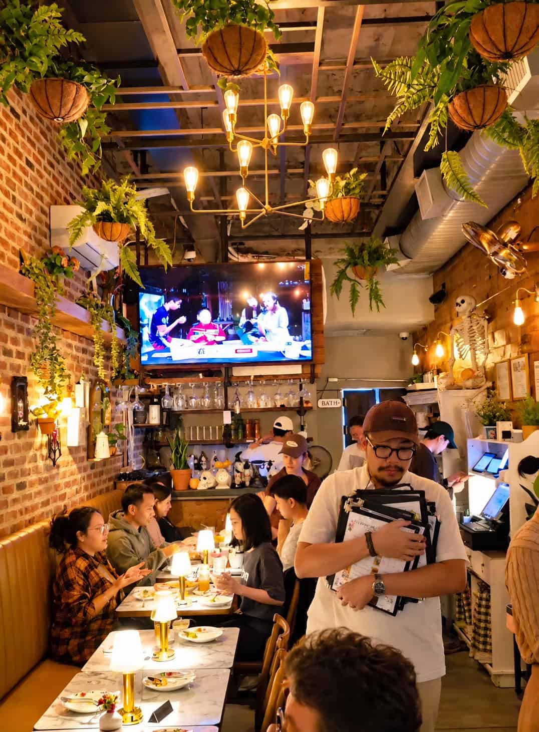 Packed dining room at SaRanRom Thai with wooden walls brick accents hanging plants and staff carrying menus