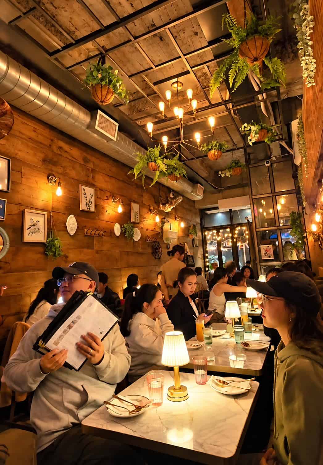 Diners seated at marble tables under warm lighting and hanging plants at SaRanRom Thai in Elmhurst