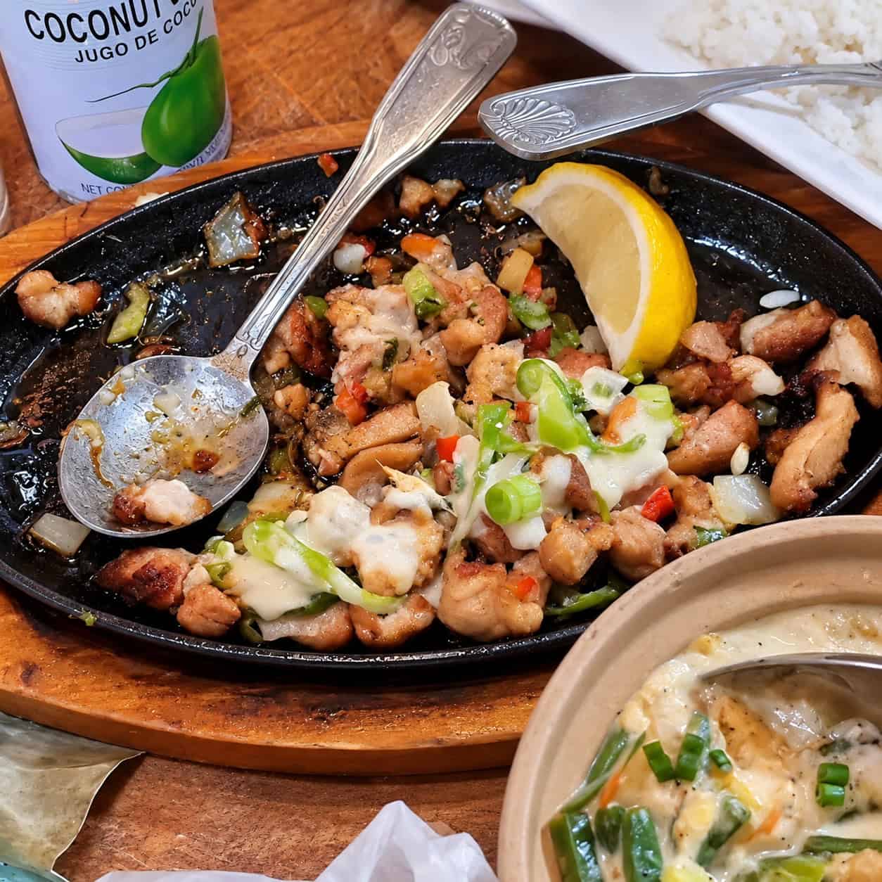 Sizzling cast-iron plate of Filipino Chicken Sisig with chopped chicken, peppers, onions, and lemon wedge