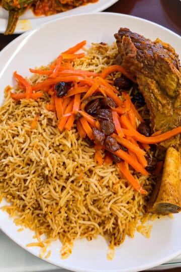 Platter of Uzbeki Qabuli Pulao featuring a large tender lamb shank over spiced brown rice topped with shredded carrots and raisins