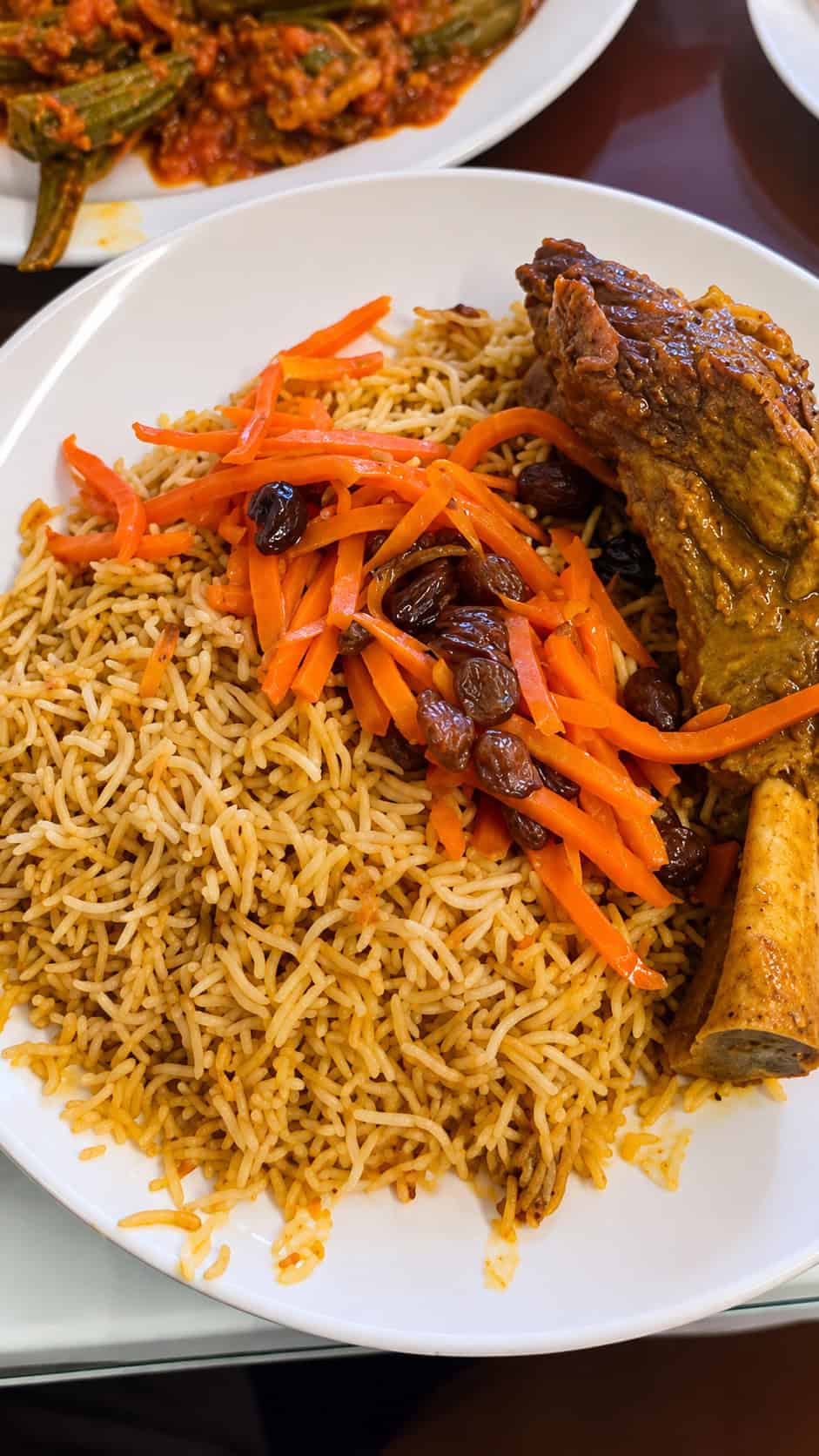 Platter of Uzbeki Qabuli Pulao featuring a large tender lamb shank over spiced brown rice topped with shredded carrots and raisins